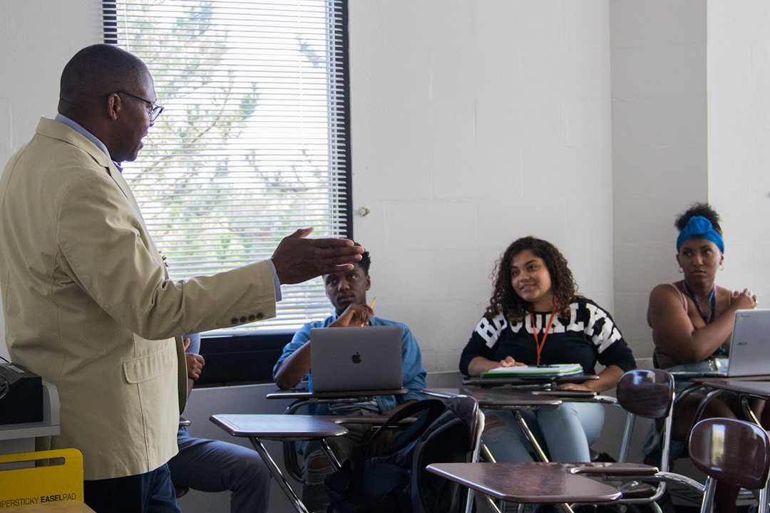 Keith Jenkins stands at the front of a room and speaks to three students who sit at desks with their laptops and paper for notes.