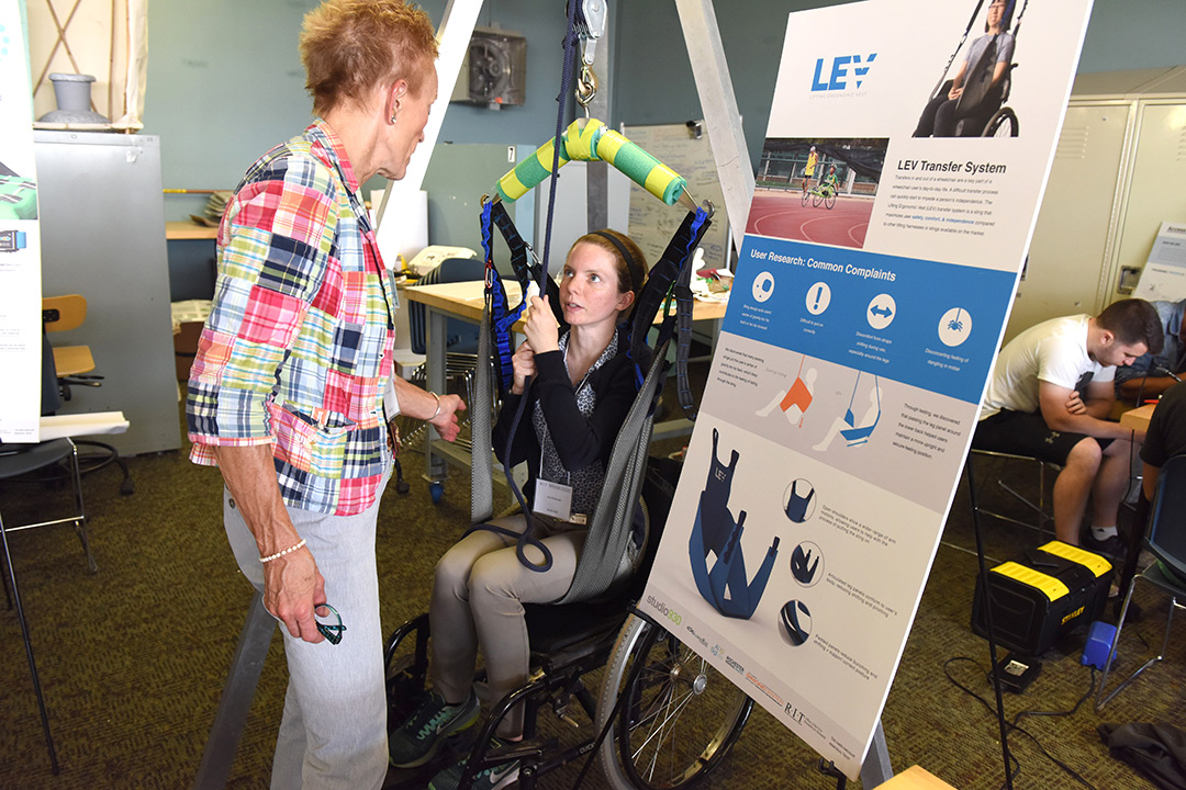 Ana Sorensen sits in a wheelchair and demonstrates how the LEV lift works for individuals to lift themselves up. She shows this to another woman who is engaged in the demonstration and reaches out to touch the assistive device.