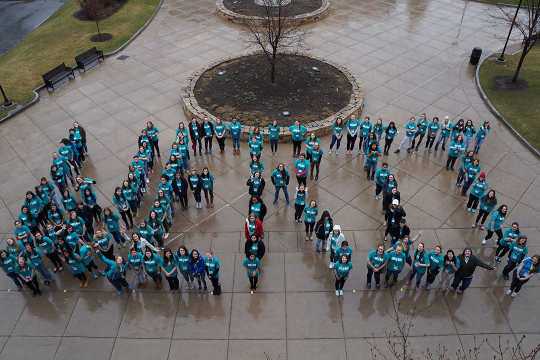 A group of WE@RIT students stand outside in their WE@RIT shirts, using their bodies to spell out "WE RIT." The photo is taken from above.