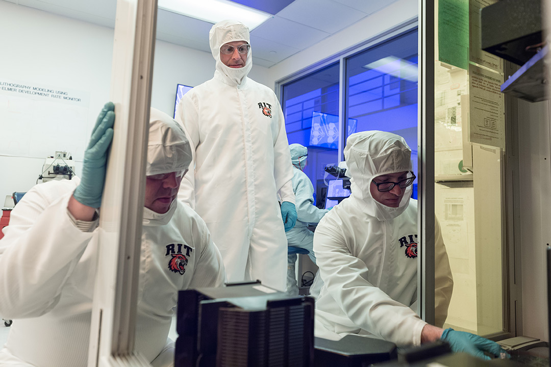 Three researchers dressed in white, sterile jumpsuits work together in a lab.