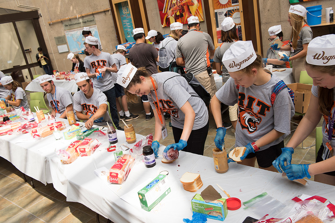 Students form an assembly line making peanut butter and jelly sandwiches. All of them are wearing matching RIT shirts and paper Wegmans hats.