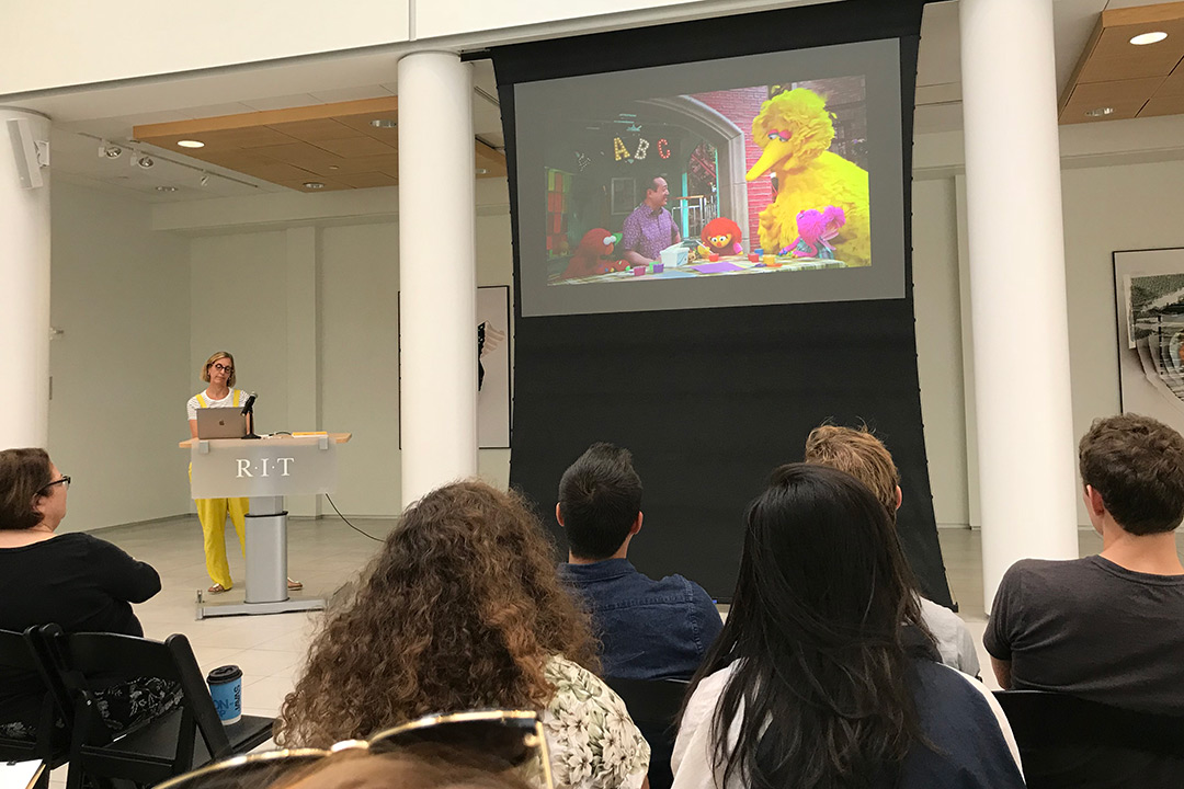 A woman in a yellow and white jumpsuit stands at a podium in front of a crowd speaking. There is a large projection screen showing a clip of Sesame Street.