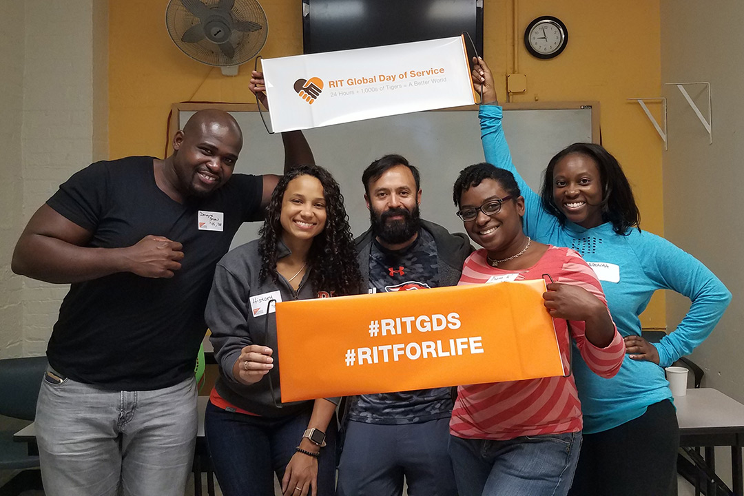 A group of alumni pose for a photo holding two signs, one reading "#RITGDS #RITFORLIFE" and the other reading "RIT Global Day of Service."