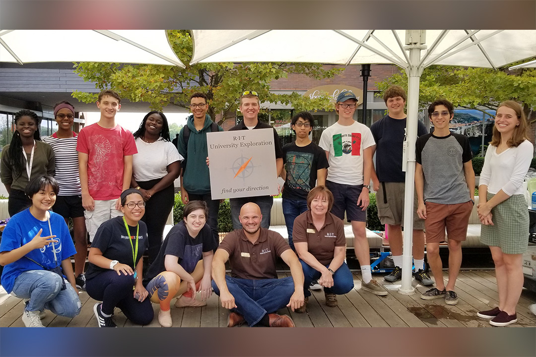 A group of University Exploration students stand together to pose for a photo, holding a sign that reads "RIT University Exploration, find your direction."