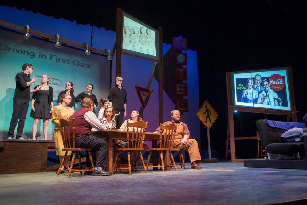 An action photo of the cast performing the show "How I learned to Drive." Six people sit and stand around a dinner table while five others dressed in black stand on a raised platform behind them.