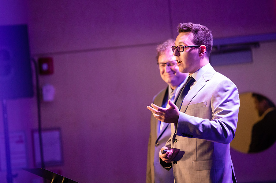 Brandon Hudson stands in a suit with purple lighting on him, speaking about his business at the competition.