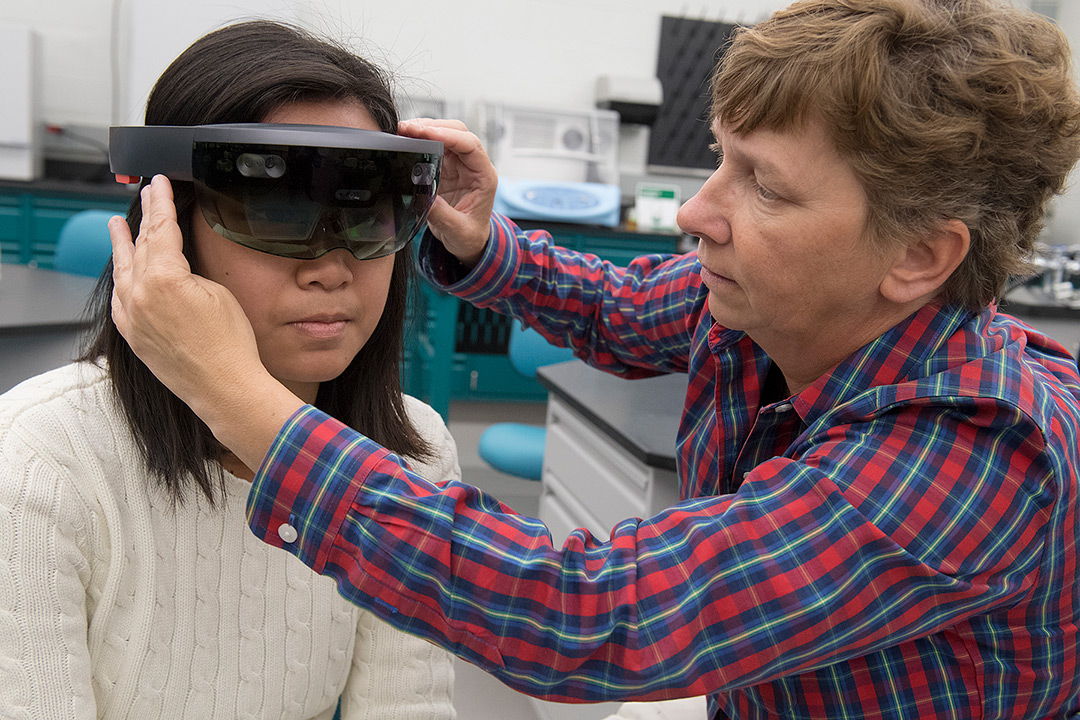 One woman on the right adjusts augmented reality glasses on another woman who is wearing them on the left.