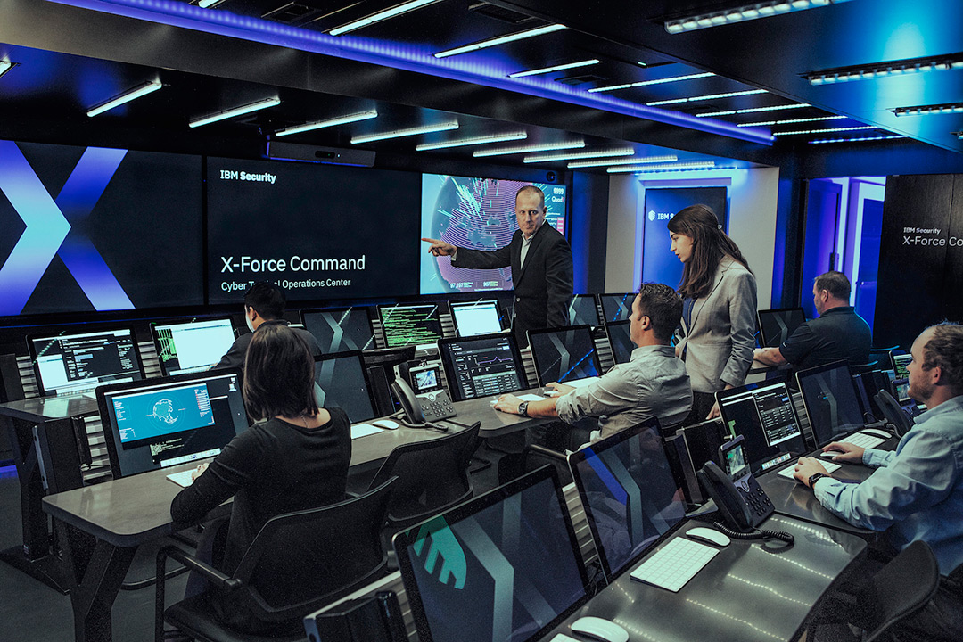 Workers sitting in front of computers and discussing what is displayed on the main screen at the front of the room. One man is pointing to the front screen.