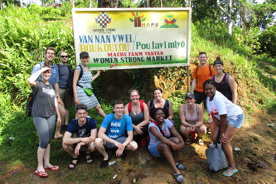 A group of students standing in front of a "Women Strong Market" sign. Most of the  words on the sign are in Haitian.