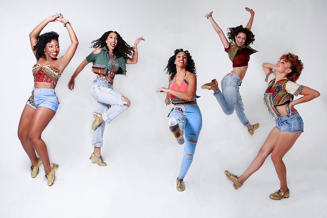 Five women dressed in jeans, colorful clothing and tap shoes jump in the air.