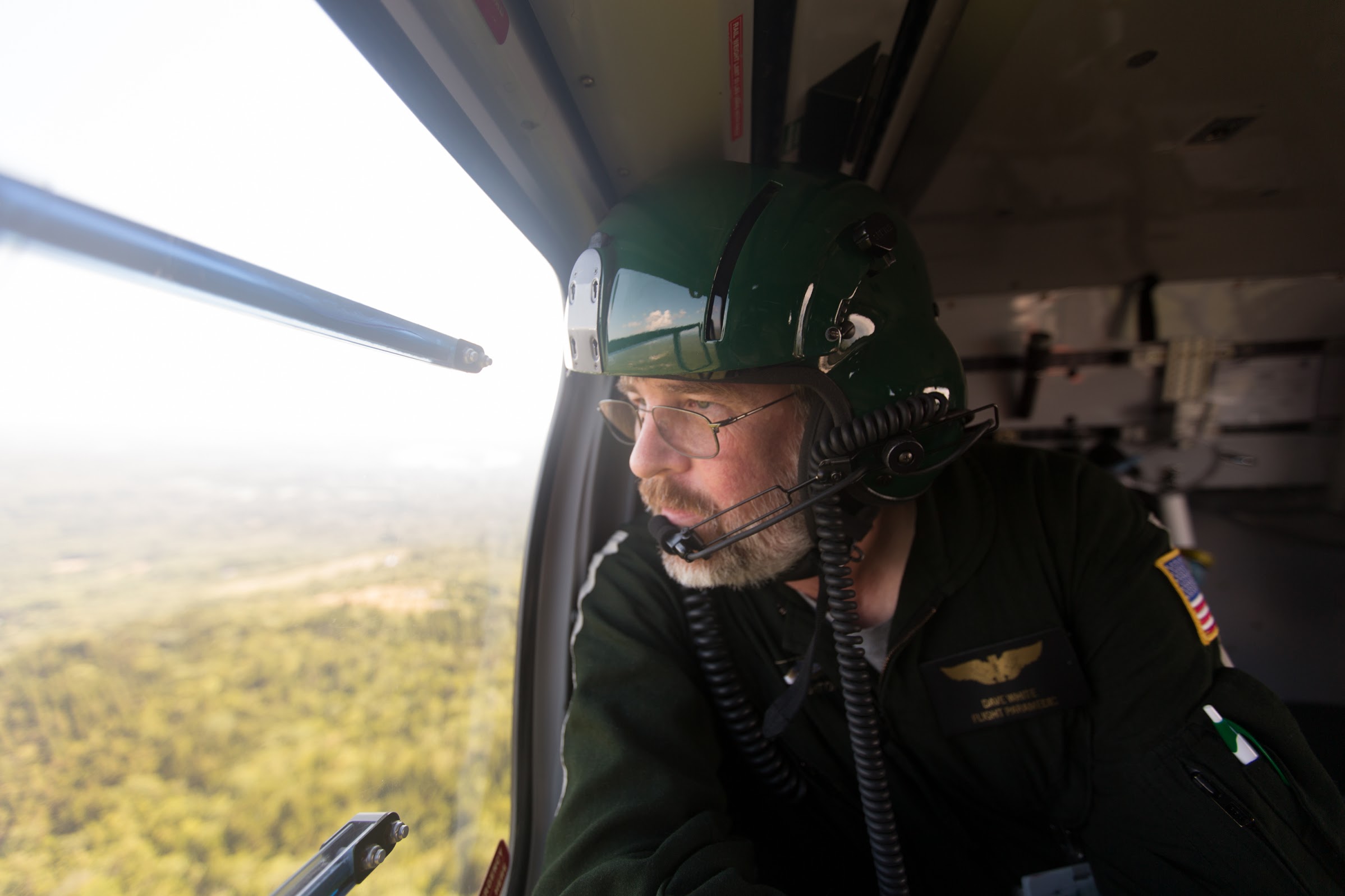 Man with pilot helmet and aviator suit looks out of a helicopter window