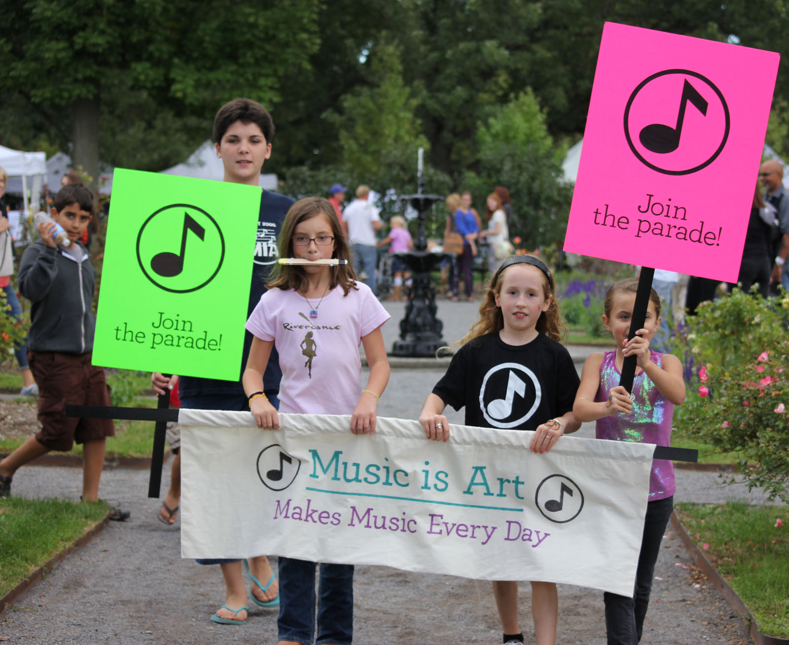 Community members hold a banner saying "Music is Art" with the subtext "Makes music every day". Students flanking the banner hold sings with the text "Join the parade"