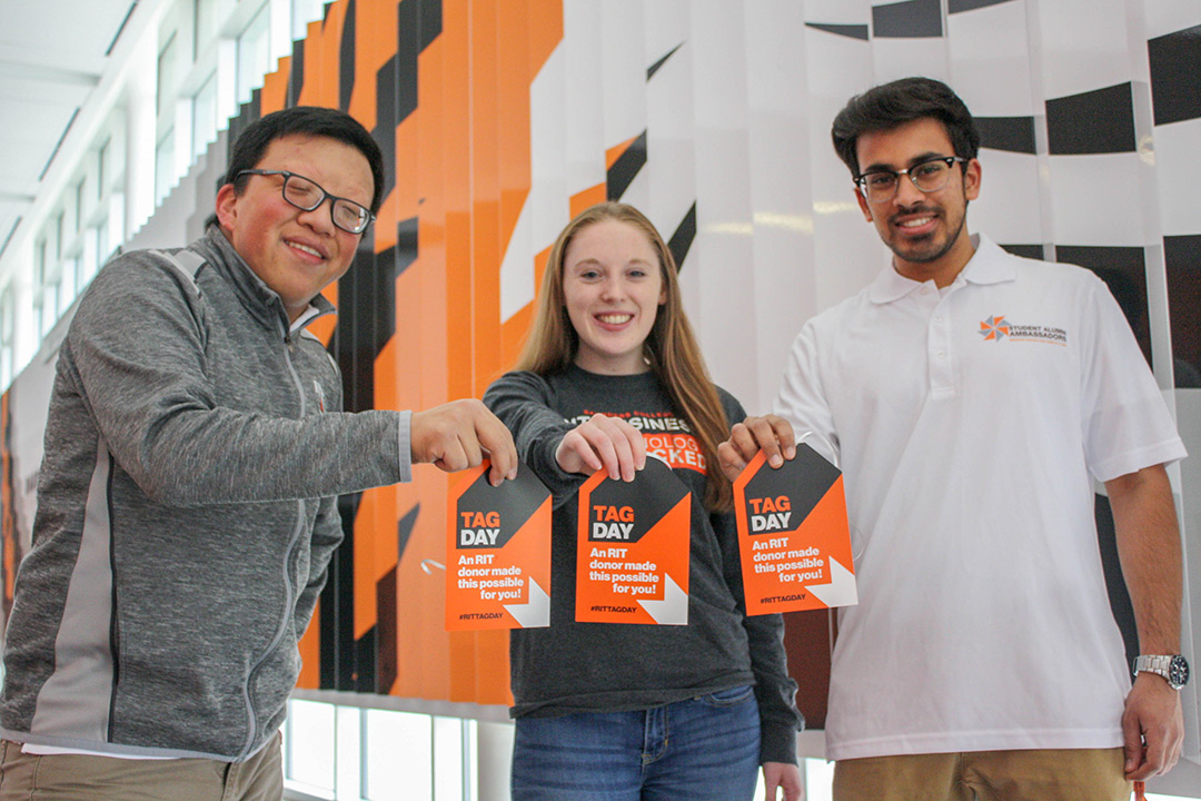 Three student stand holding orange tags that read: "TAG Day: An RIT donor made this possible for you."