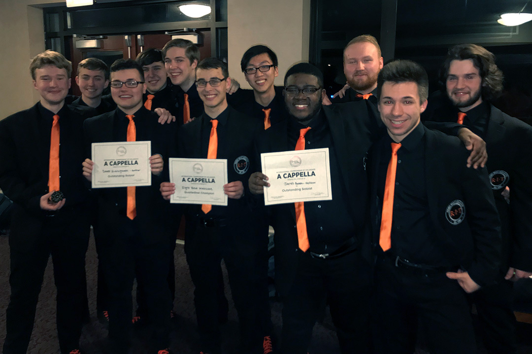 Group of 11 students in black suits and orange ties stand, with three students holding certificates