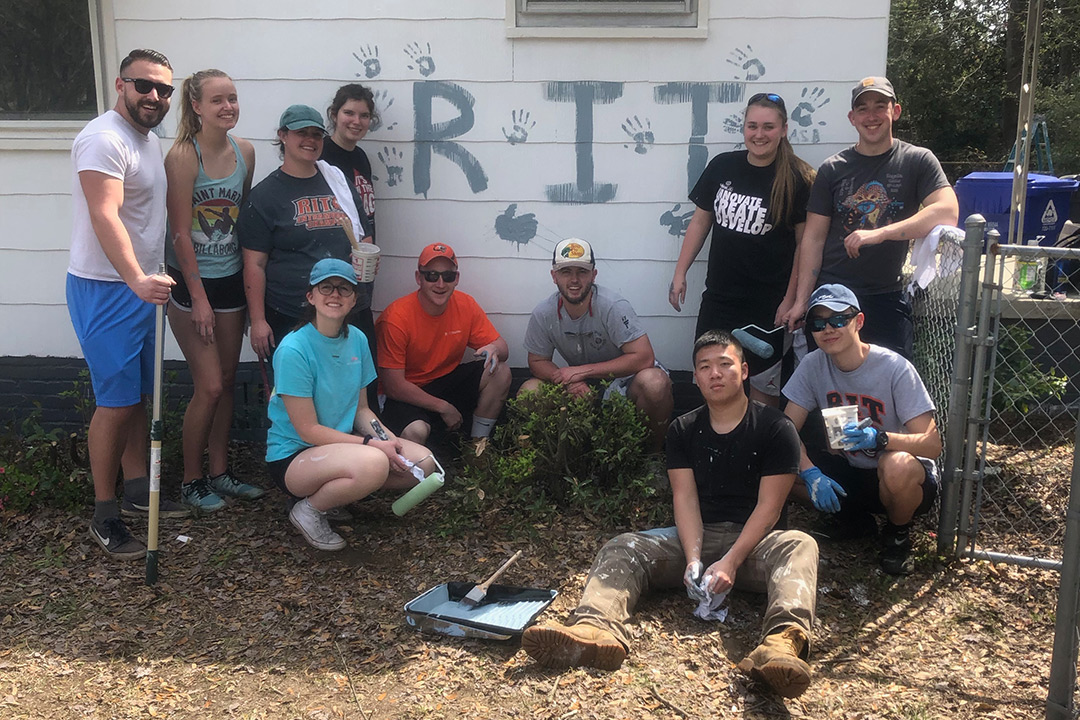 Group of students poses around outside of a house they are painting, with the letters RIT and their handprints painted on the house exterior.