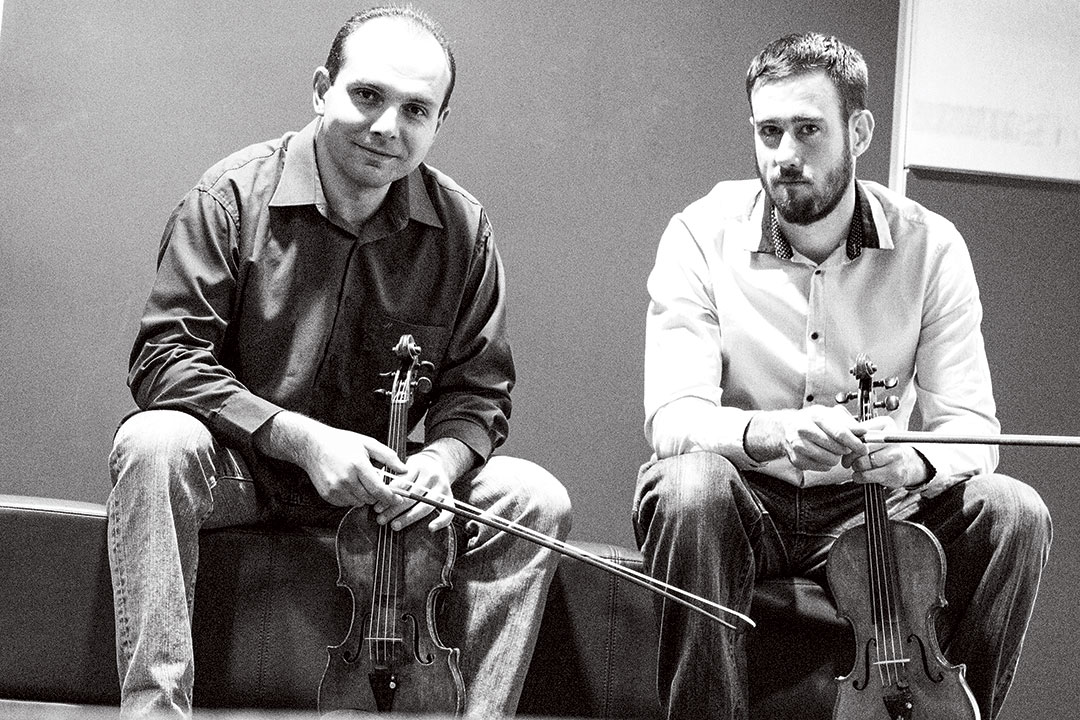 Two men sit holding violins.