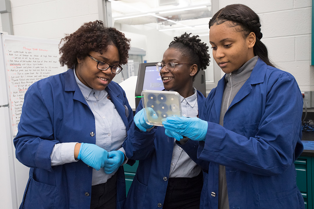 Three high school students wearing blue lab coats examine petri dish.