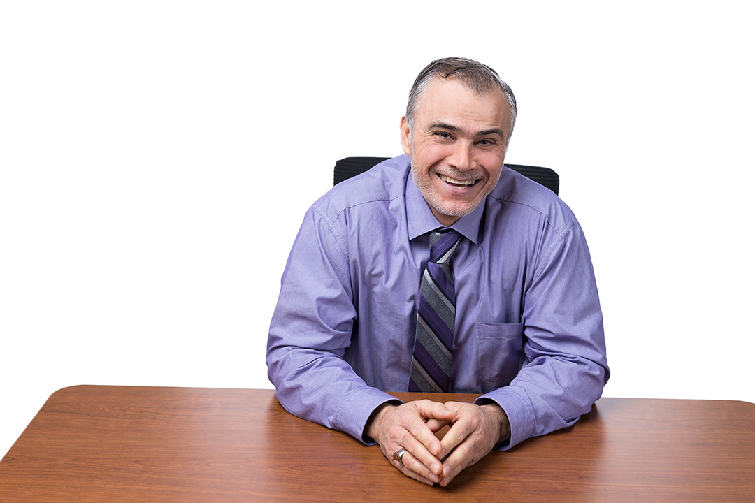 Man wearing purple button-up shirt and tie sits at desk.