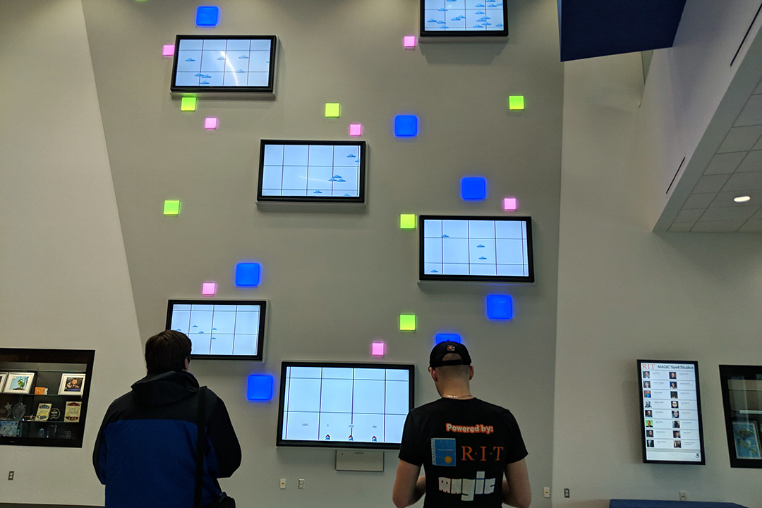 Two students play video game on six TVs mounted to wall.