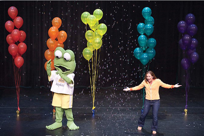 Woman performs on stage with large frog mascot.