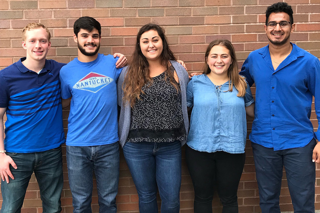 Group of five students stands against a brick wall.