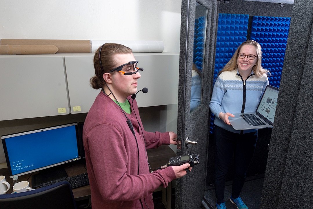 Student wearing eye-tracking headset stands with another student holding laptop.