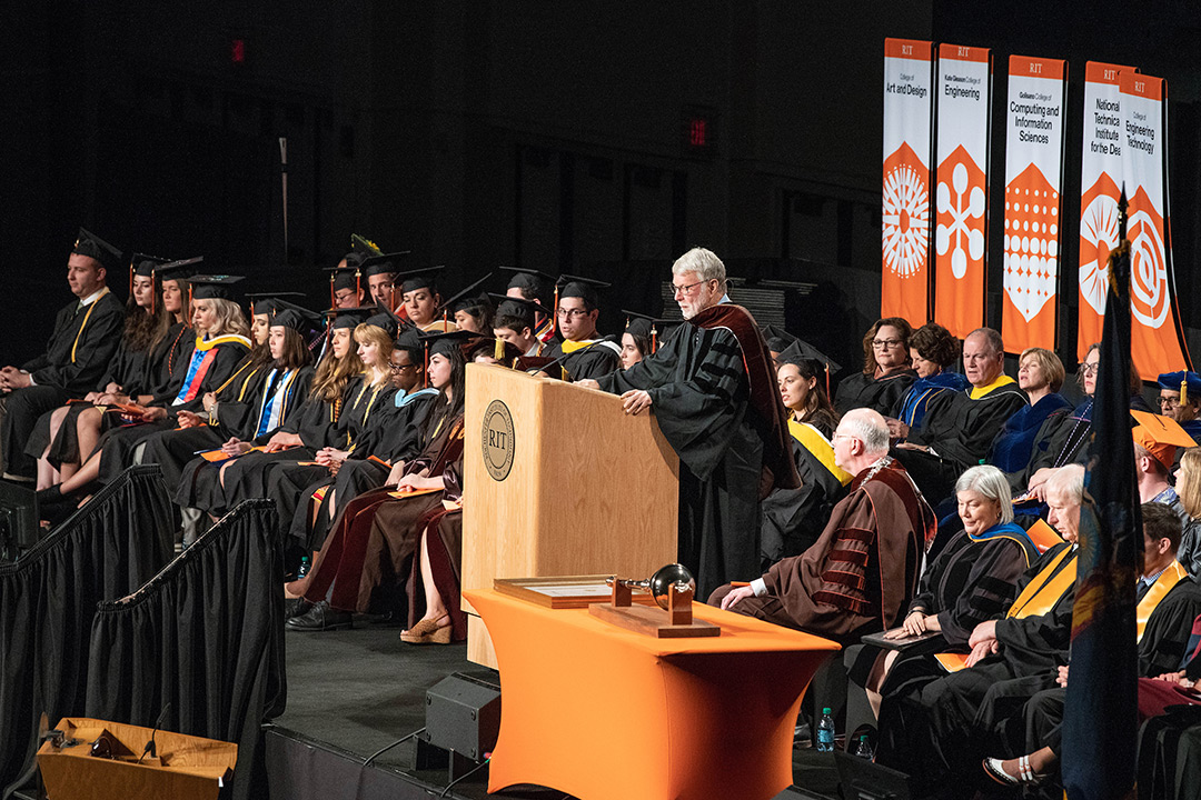 Man in commencement regalia speaks at podium on stage.