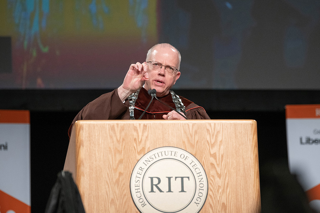 Man in commencement regalia speaks at podium on stage.