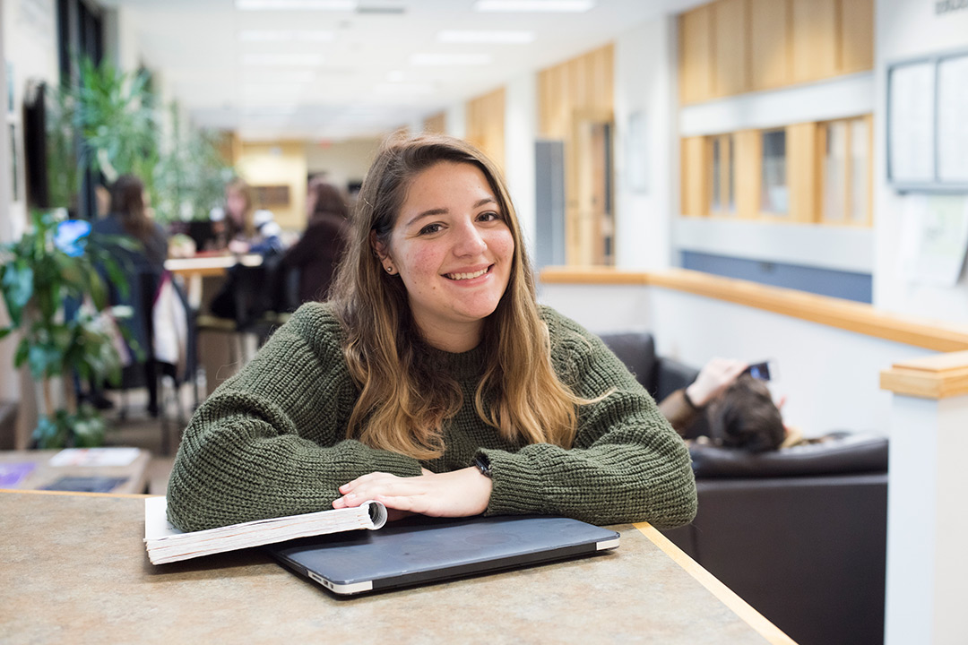 Student poses sitting at table with notebook and laptop.