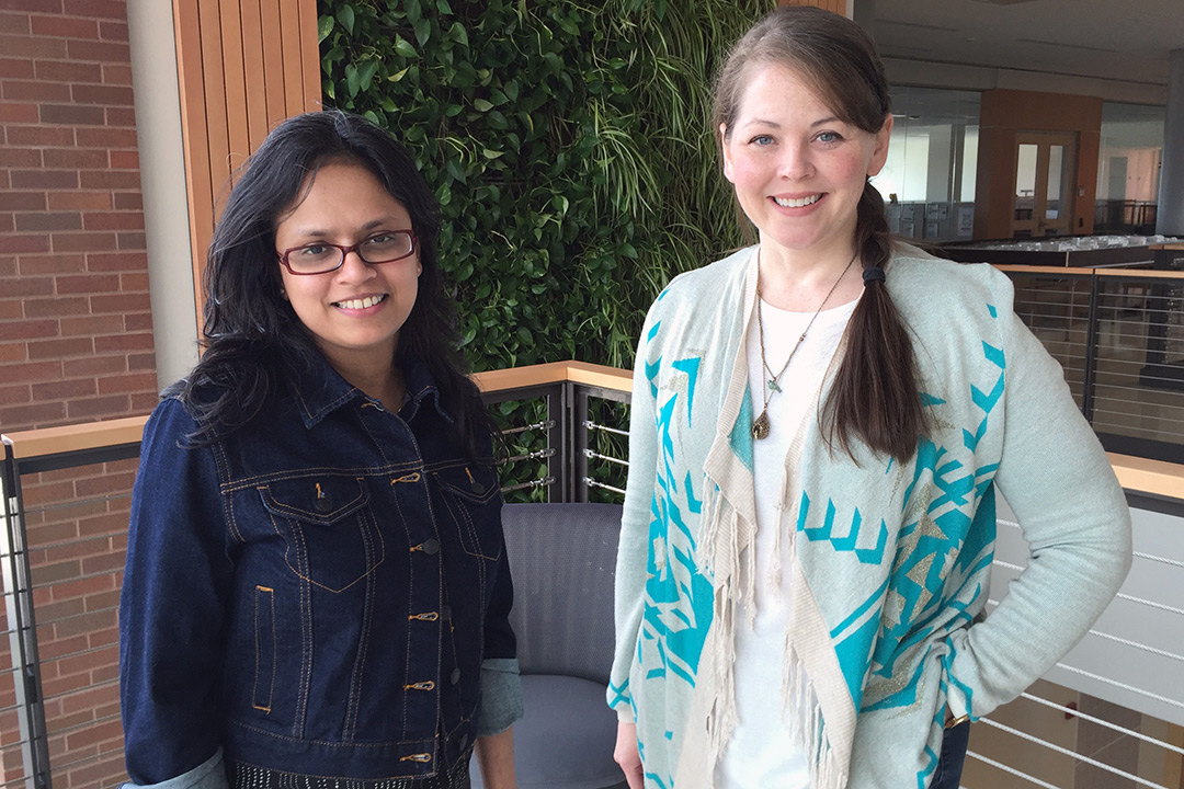 Two women stand in front of wall with plants.