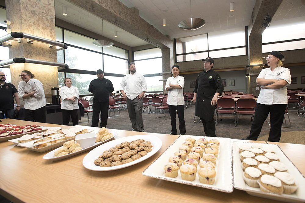 Libe of chefs stands behind table with plates of desserts.