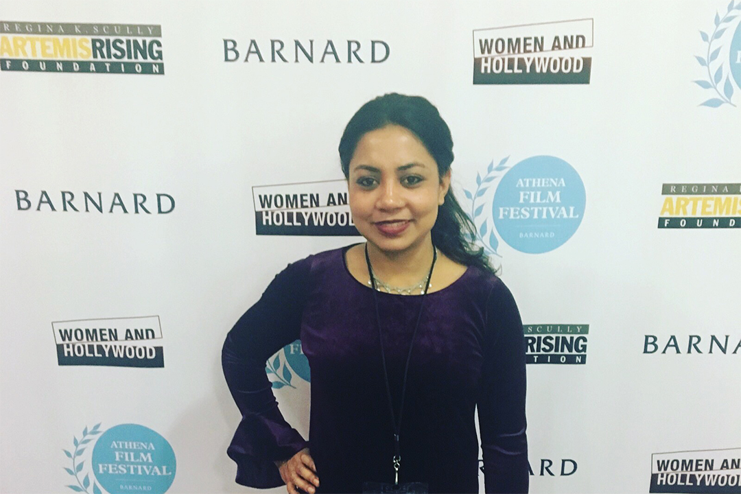 Woman in purple shirt standing in front of white background with logos.