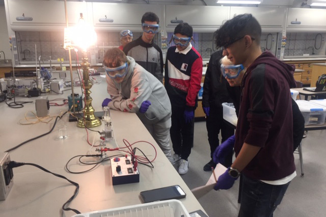 Students wearing eye protection conduct experiment with a multimeter and lamp.