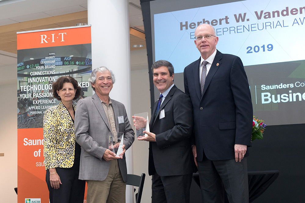 Four people stand together, with two in center holding glass awards.