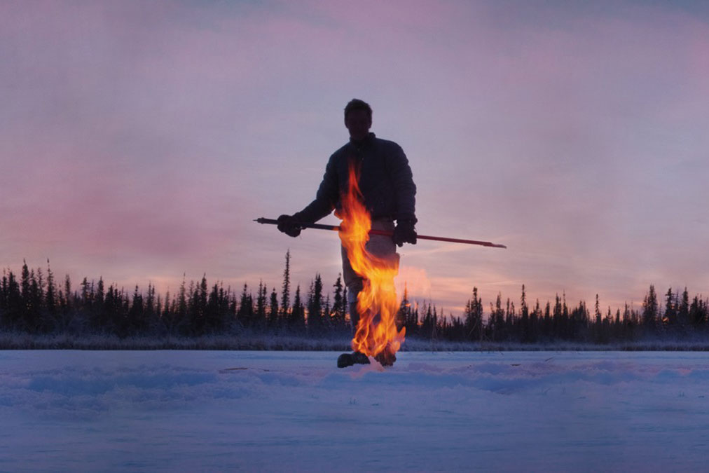 Person walks across snow as flame shoots up from pipe in the snow.