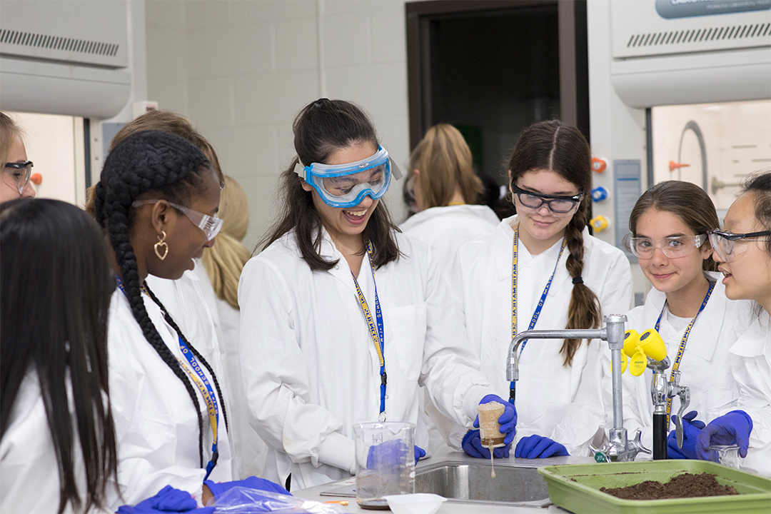 Girls in lab coats wearing safety goggles conducting an experiment over a sink.