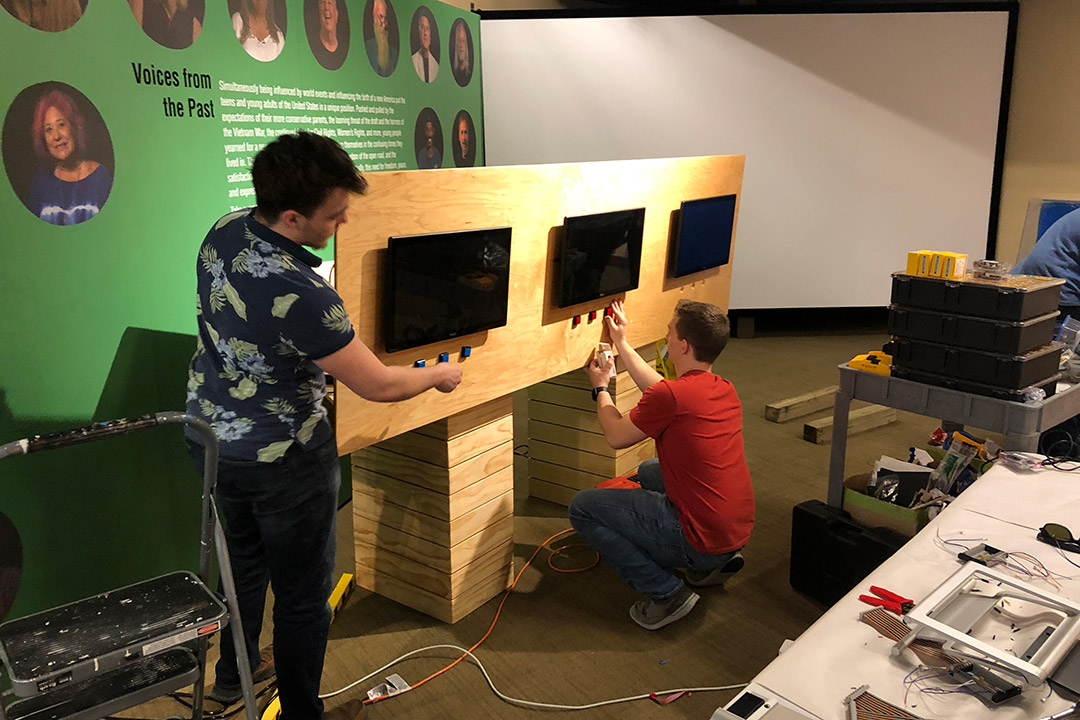 Students set up kiosk with three TV monitors and buttons underneath.