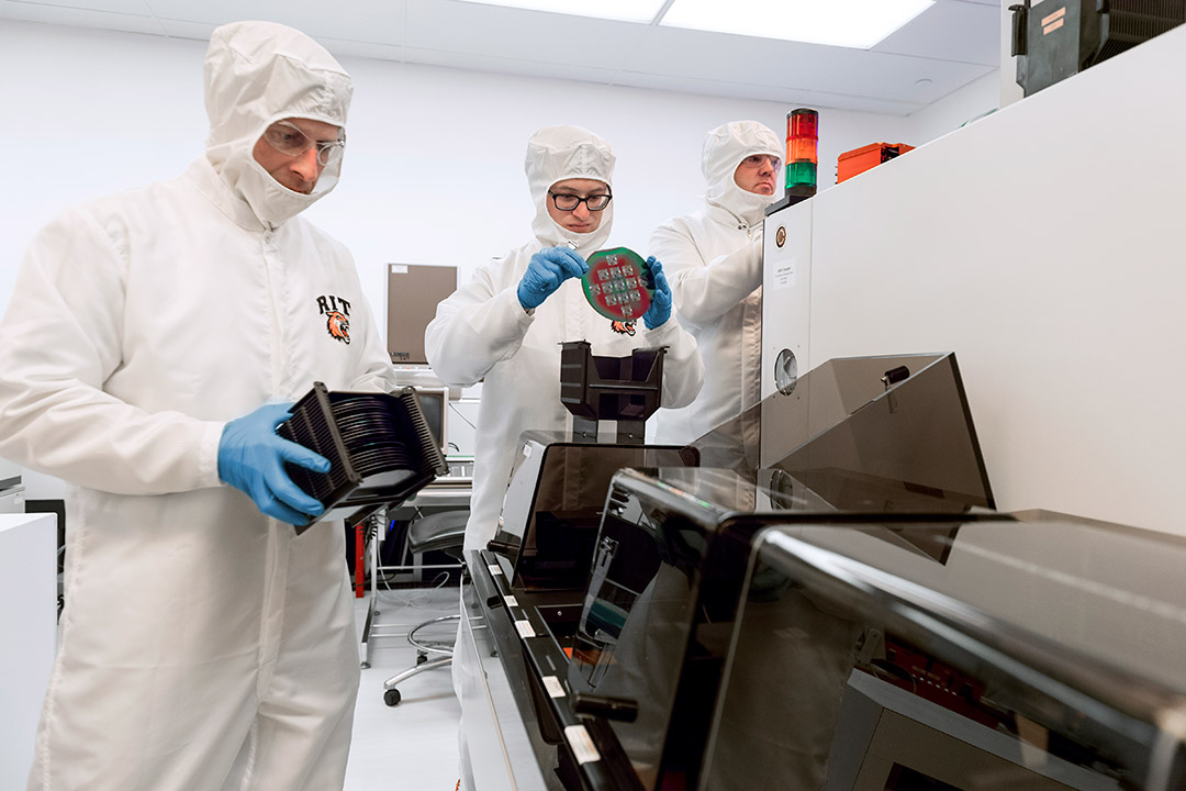 Researchers wearing white cleanroom suits handle circuits.