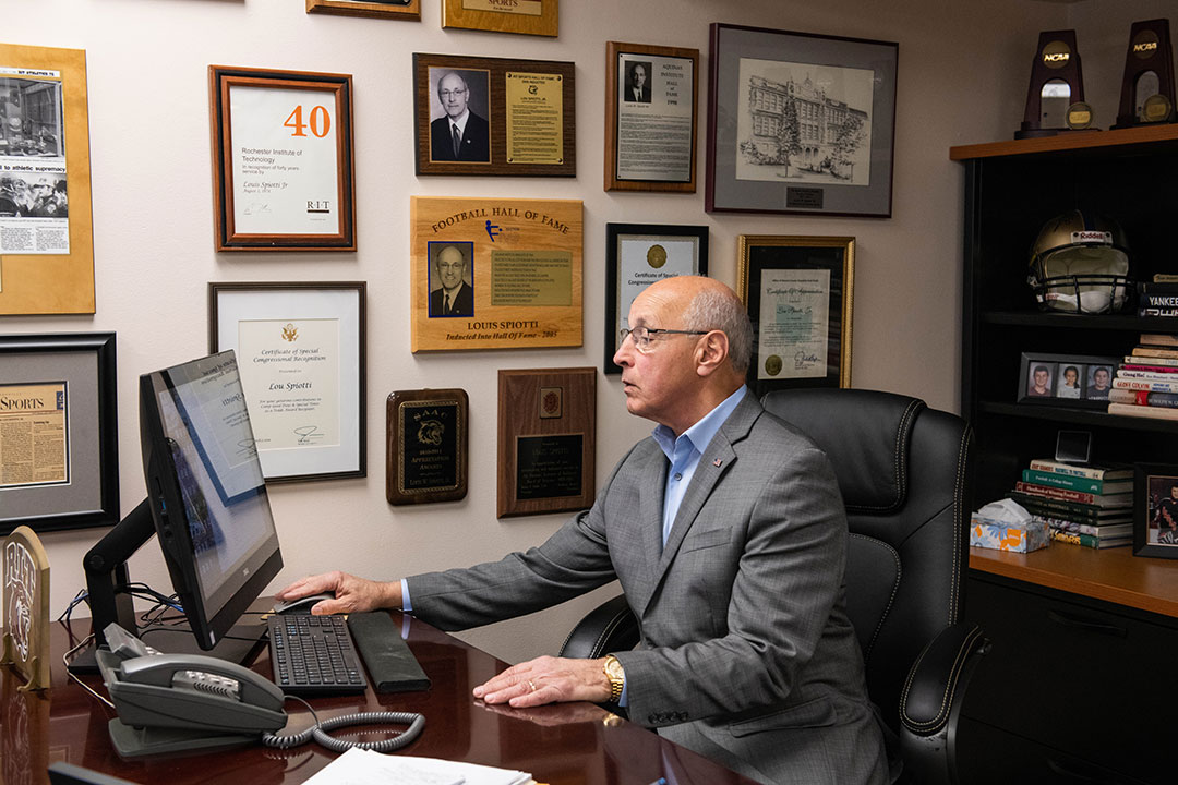 Man wearing suit sits at office desk with awards on wall behind him.