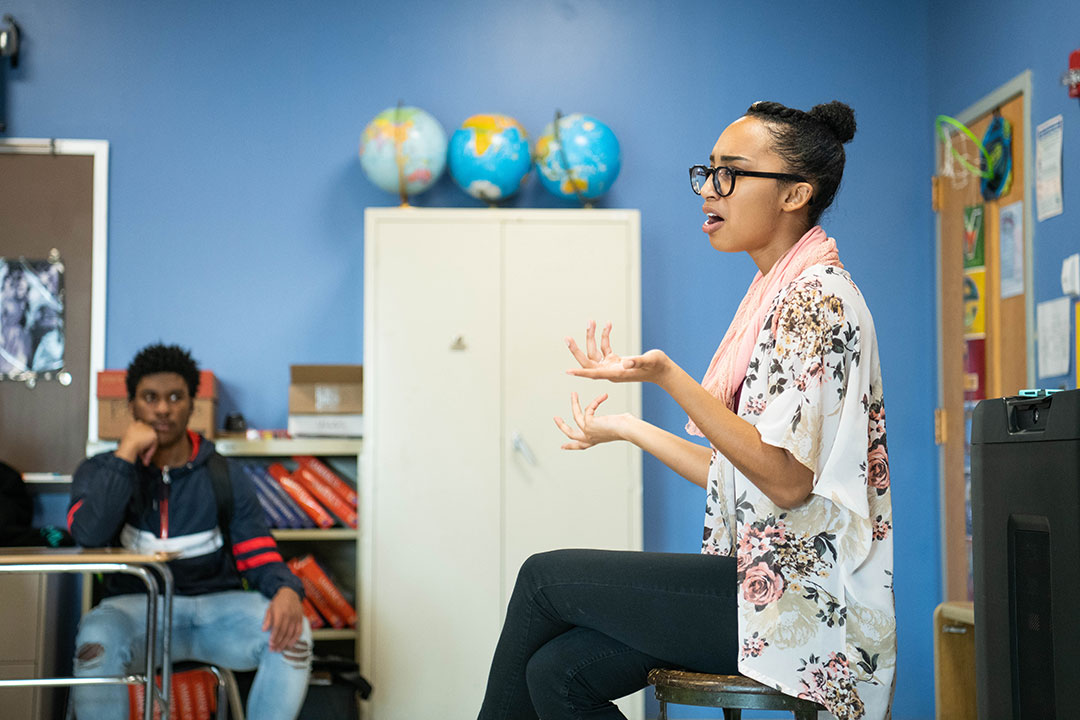 Woman sits on stool speaking to classroom.