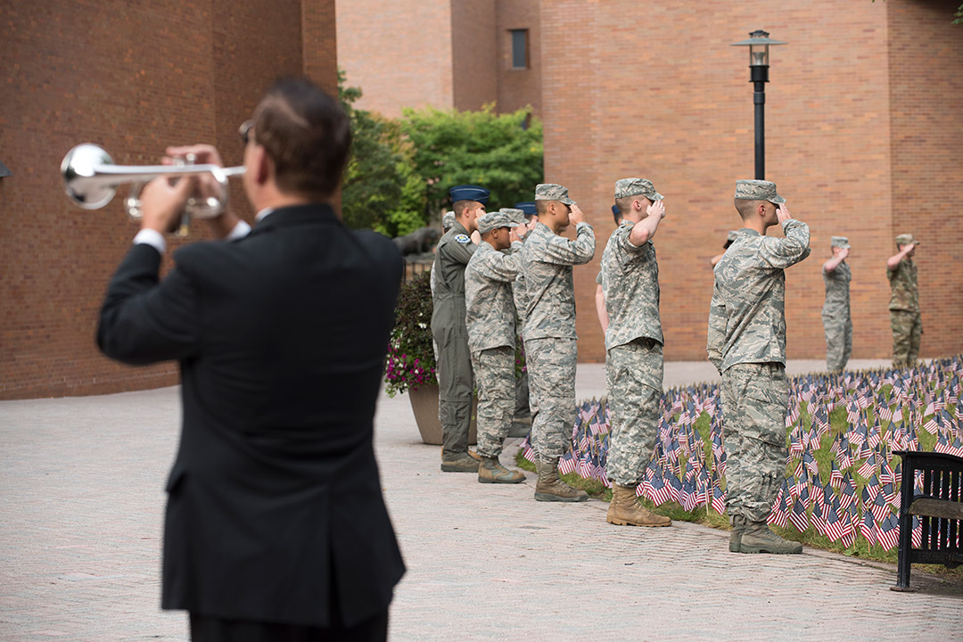 Man plays Taps while ROTC members salute field of American flags.