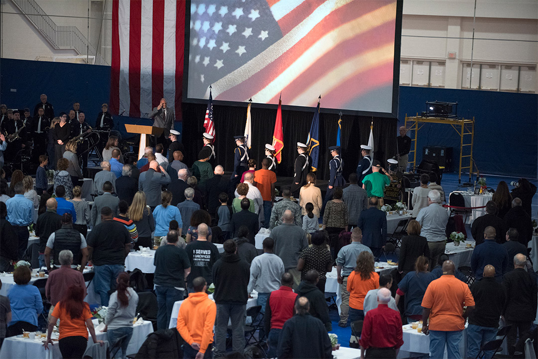 Overhead view of group of people at several tables, standing in honor of uniformed military members.