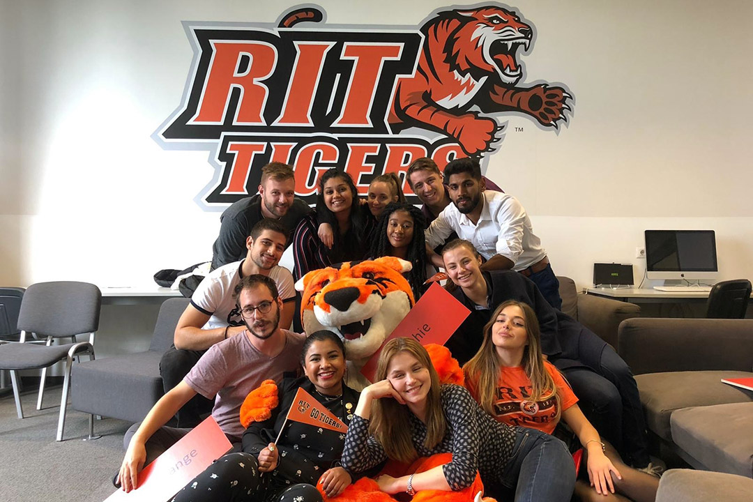 Students pose wth tiger mascot in front of RIT Tigers logo on wall.