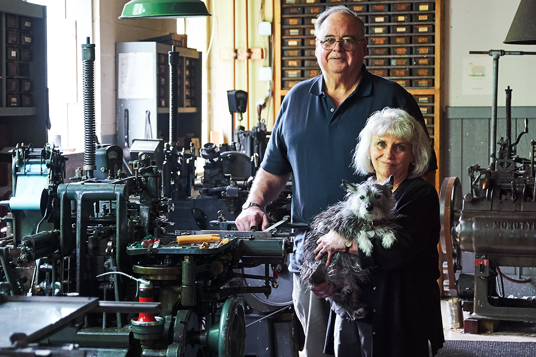 Man and woman holding dog stand next to printing presses.