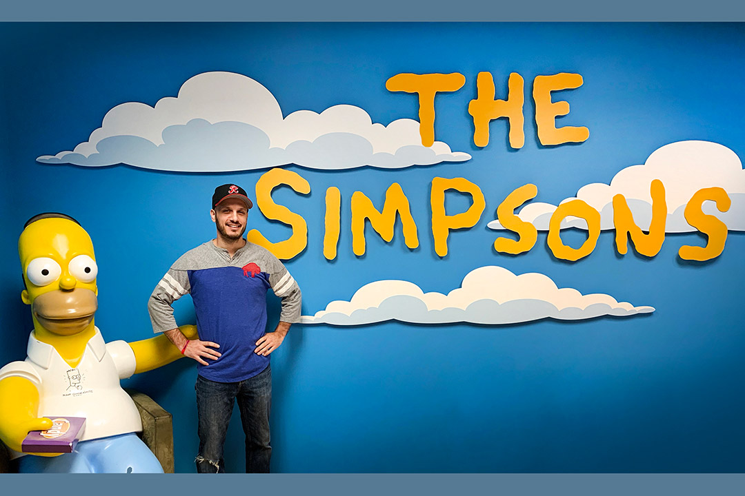 Man stands next to statue of Homer Simpson in front of iconic opening scene of 'The Simpons' title appear on clouds in blue sky.