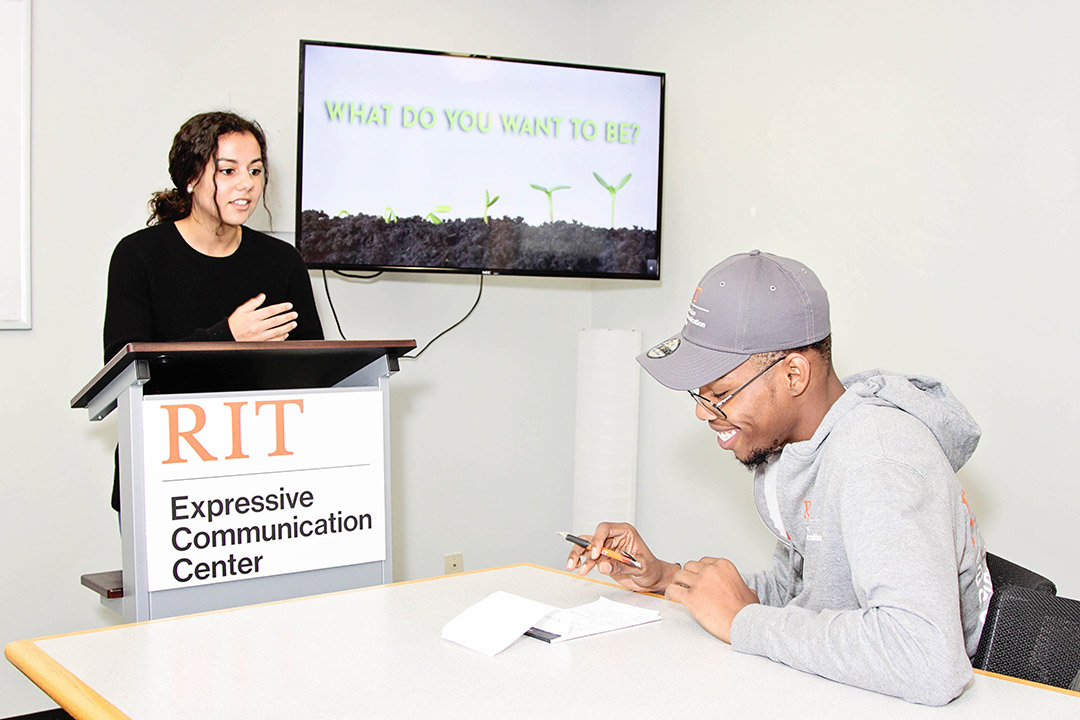 student standing at podium and another sitting at a table with a TV screen displaying: What do you want to be?