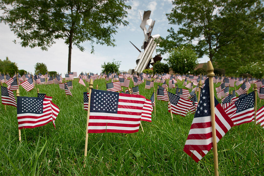 small American flags stuck into the lawn in front of a metal sculpture.