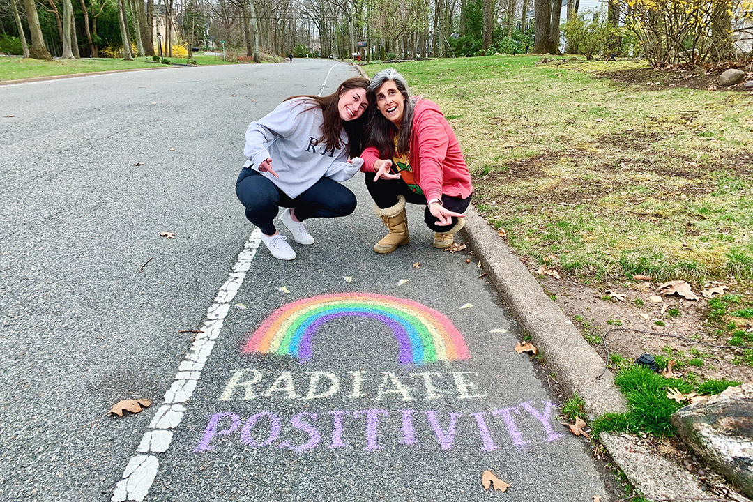 mother and daughter posing on street above sidewalk chalk art of the words "radiate positivity" and a rainbow.