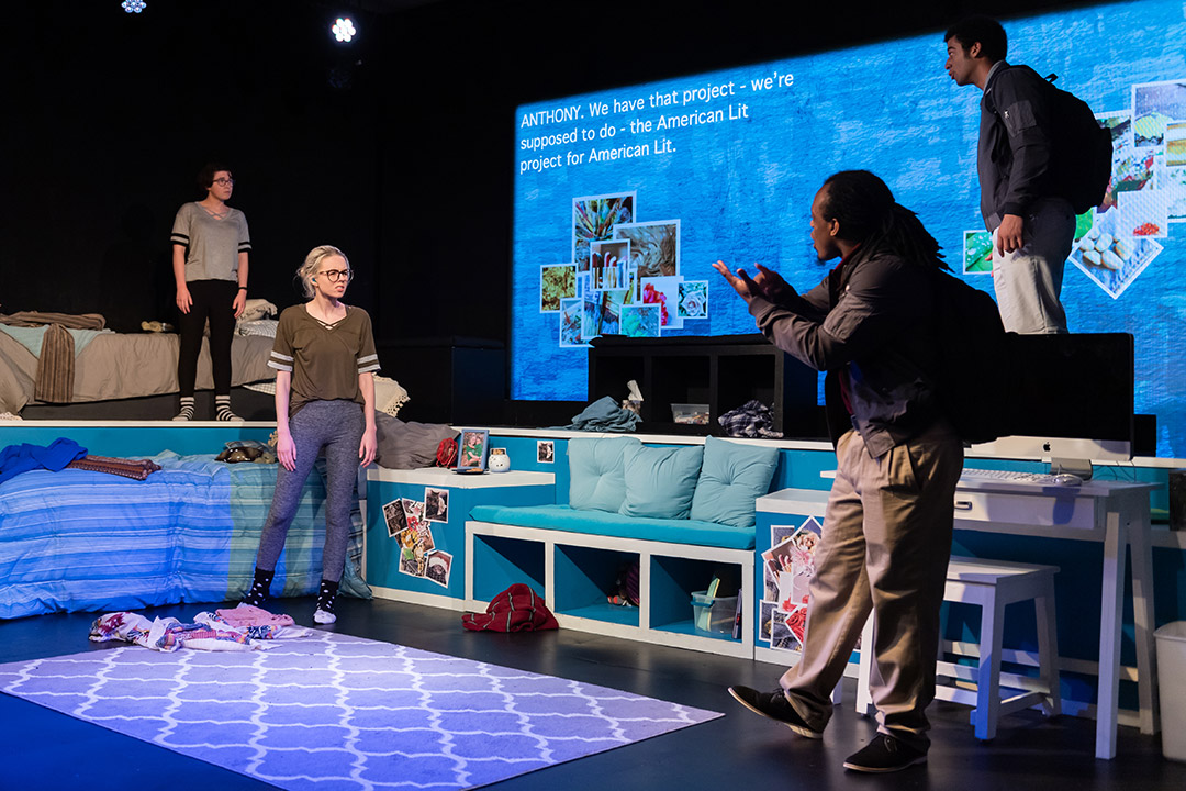 students performing a play in a set that looks like a college dorm room.