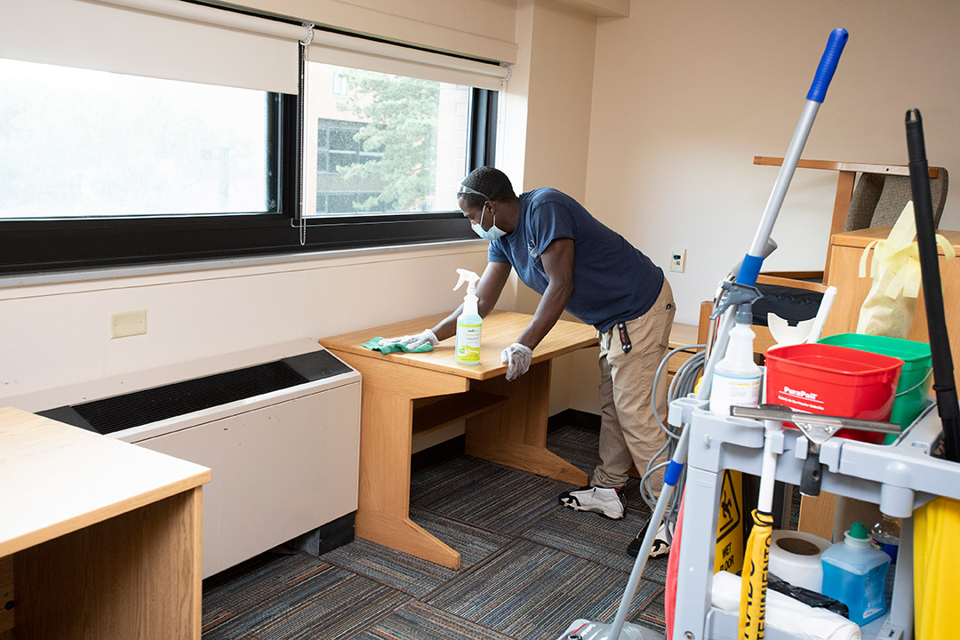 man wearing face masks cleaning a desk in a dorm room.